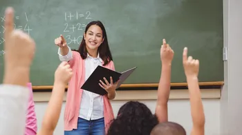Lehrerin vor der Tafel zeigt dynamisch in die Klasse, im Fordergrund zur Meldung hochgestreckte Finger der Schüler*innen